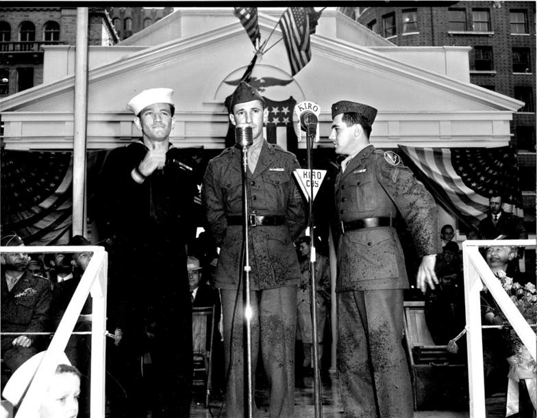two Marines and one Navy on a stage in uniforms in front of a microphone