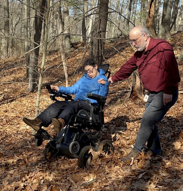 girl in blue shirt in an iBot motorized wheelchair is guided by an older man in red shirt as they go through a trail in the woods on a fall day.
